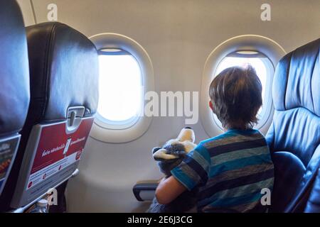 Ein Kind in einem Flugzeug hält ein weiches Spielzeug und Tragen einer Gesichtsmaske, die aus dem Fenster schaut Stockfoto