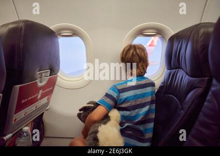 Ein Kind in einem Flugzeug hält ein weiches Spielzeug und Tragen einer Gesichtsmaske, die aus dem Fenster schaut Stockfoto