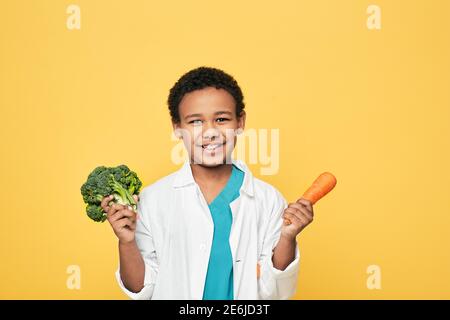 Ein lächelnder männlicher Junge hält Karotten und Brokkoli in der Hand. Gesundes vegetarisches Essen für Kinder. Gelber Hintergrund Stockfoto