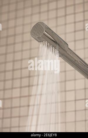 Nahaufnahme des modernen Edelstahl-Duschkopfes mit Wasser, das im Hintergrund mit Fliesen im Badezimmer fließt. Low-Angle-Ansicht und selektiver Fokus. Stockfoto