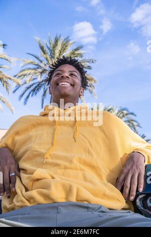 afro amerikanischer schwarzer Mann mit gelbem Sweatshirt beim Blick auf Smartphone Auf einer Holzbank im Park liegend Stockfoto