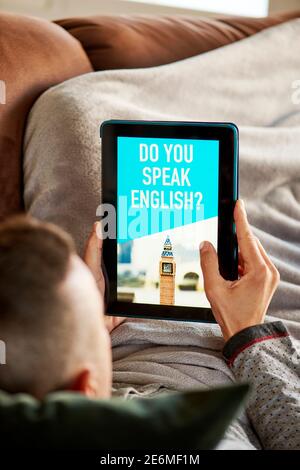 Ein junger Kaukasusmann sitzt bequem in einem Sofa, das in eine wärmende graue Decke gehüllt ist, und hat sein Tablet in den Händen, mit dem Text sprichst du Stockfoto