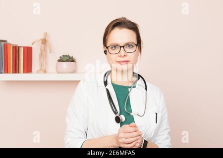 Portrait von selbstbewussten jungen Arzt trägt weißen Mantel mit Stethoskop und Hörmuschel, die in ihrem Home Office stehen und sie betrachten Kamera bereit für onl Stockfoto