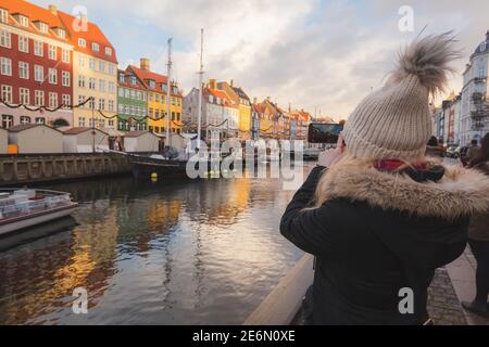 Eine junge blonde Touristin im Winter macht ein Foto von goldenem Sonnenlicht an der farbenfrohen Uferpromenade des Nyhavn Canal (New Harbour), einem Unterhaltungs-d Stockfoto