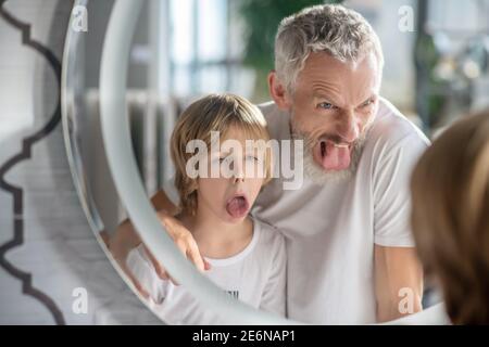 Mann und sein Sohn machen lustige Gesichter in der Nähe des Spiegels Stockfoto