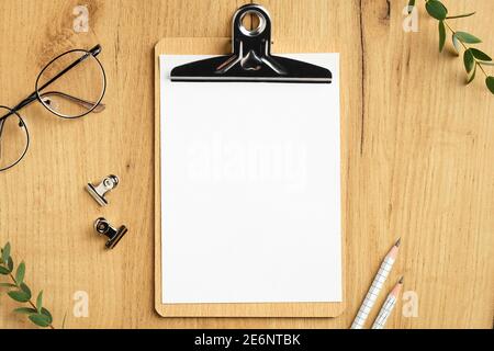 Holzschreibtisch mit leerem Papier Clipboard Mockup, Gläser, Bürobedarf, Eukalyptus Blätter. Flach liegend, Draufsicht. Stockfoto