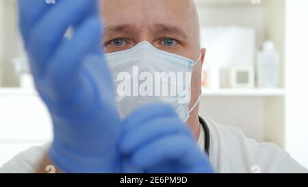 Arzt trägt eine schützende Gesichtsmaske und legt OP-Handschuhe auf seine Hände. Stockfoto
