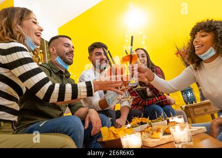 Gruppe junger gemischter Renner feiern zu Hause tragen medizinische Schutzmaske sitzen auf dem Sofa um beladenen Tisch. Happy Hour toasten zu Hause mit Stockfoto