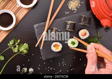 Hände mit Essstäbchen pflücken Sushi aus Schieferteller mit Sortiment auf dem Tisch mit Zutaten und Behälter und dunklen Hintergrund. Draufsicht. Horizonta Stockfoto