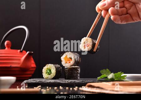Hände mit Essstäbchen pflücken Sushi aus Schieferteller mit Sortiment auf dem Tisch mit Zutaten und Behälter und dunklen Hintergrund. Vorderansicht. Horizont Stockfoto