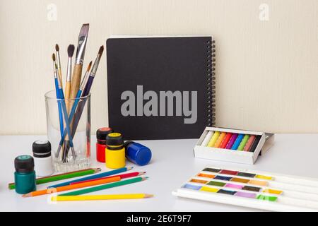 Ein schwarzes Skizzenbuch auf einem weißen Tisch mit Zeichenmaterial Und Bürsten Stockfoto