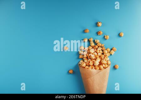 Karamell Popcorn in einem Papierumschlag auf blauem Hintergrund. Köstliches Lob für das Ansehen von Filmen, Serien, Cartoon. Freier Speicherplatz, Nahaufnahme. Minimalis Stockfoto