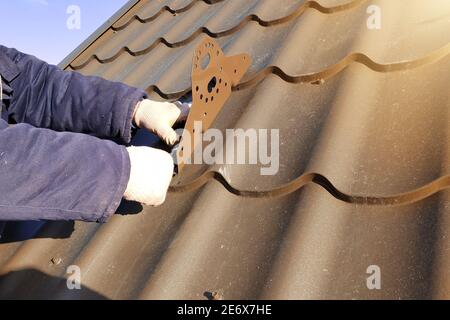 Der Arbeiter arbeitet in der Höhe, befestigt die Beschneiungselemente auf dem verzinkten Dach, arbeitet in der kalten Jahreszeit, der Arbeiter arbeitet ohne Versicherung. 2020 Stockfoto