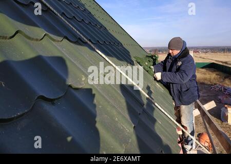 Der Meister arbeitet auf dem Dach eines Privathauses ohne Versicherung, die Arbeit wird in der kalten Jahreszeit durchgeführt, der Arbeiter verhält sich vorsichtig, das Dach ist Stockfoto