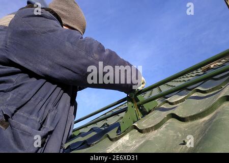 Der Arbeiter hält in den Händen die Elemente zum Dach, das Dach des Daches aus grünem Metall, der Arbeiter in Winterkleidung, arbeitet in der Höhe ohne ins Stockfoto