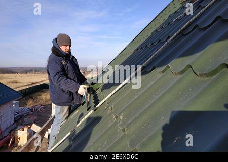 Der Arbeiter hält in den Händen die Elemente zum Dach, das Dach des Daches aus grünem Metall, der Arbeiter in Winterkleidung, arbeitet in der Höhe ohne ins Stockfoto