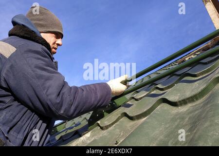 Der Arbeiter hält in den Händen die Elemente zum Dach, das Dach des Daches aus grünem Metall, der Arbeiter in Winterkleidung, arbeitet in der Höhe ohne ins Stockfoto