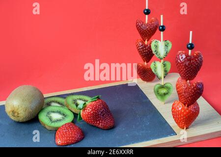 Früchte Kiwi und Erdbeeren in Herzform auf den Stäbchen auf dem Schiefertafel und auf dem roten Hintergrund als Symbol der Liebe und Valentinstag. Stockfoto