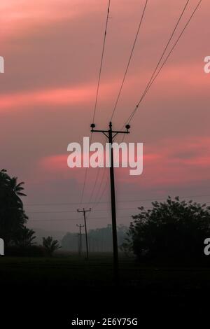 Ländliche Dorf am frühen Morgen Nebel und malerischen roten Himmel Wolken Landschaft Foto. Nicht so gerade hölzerne elektrische Pole in das Reisfeld, fährt Stockfoto