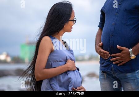 Junge attraktive schwangere Dame mit Brille Blick auf den fetten Bauch des Mannes, lustige Fotografie, paar Ziele Konzept, Outdoor-Strand Fotoshooting in G Stockfoto