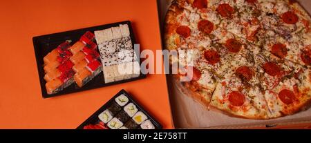 Nahaufnahme von appetitischem Sushi in Behältern und großer Pizza in Box auf orangefarbenem Hintergrund. Pizza und leckere Sushi-Brötchen mit verschiedenen Zutaten Stockfoto