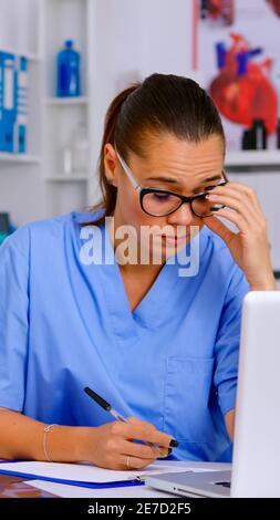 Erschöpfter Therapeut, der am Laptop arbeitet und die Brille auszieht und im Krankenhaus Kopfschmerzen hat. Verstört Therapeut Student Praxis Medizin Schreiben auf Zwischenablage Liste der diagnostizierten Patienten Stockfoto