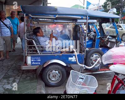 Ein Thai Tuk Tuk oder Taxifahrer ruht und Spielen mit seinem Handy während der Wartezeit auf einen Passagier während Zwei ausländische Touristen kommen hinter ihm vorbei Stockfoto