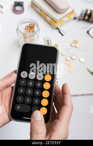 Nahaufnahme eines Taschenrechners, der von weiblichen Händen bei der Analyse aufgehängt wurde. Im Hintergrund finden sich Münzen, Banknoten und schriftliche Budgets. Stockfoto