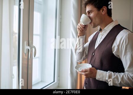 Ein junger Geschäftsmann in einem Hotelzimmer, der sich auf einen Job vorbereitet und an einem schönen Morgen einen Kaffee trinkt. Hotel, Business, Leute Stockfoto