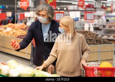 Zeitgenössische reife Paar in Casualwear und Schutzmasken Auswahl frisch Reifes Gemüse im Supermarkt, während Sie bei einem großen Display stehen Stockfoto