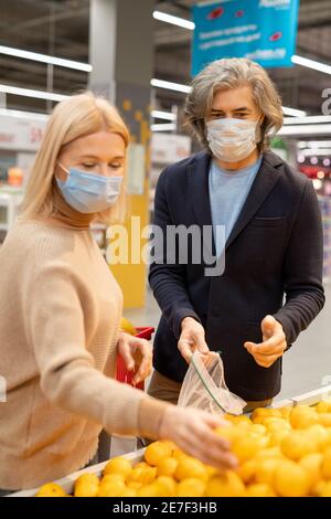 Zeitgenössische reife Paar in Casualwear und Schutzmasken Auswahl frisch Reife Früchte im Supermarkt, während man an einem großen Display steht Stockfoto