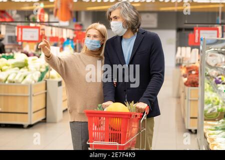 Zeitgenössische reifes Paar in Casualwear und Schutzmasken bewegen sich entlang Großer Supermarkt, während die Frau auf eine der Abteilungen zeigt Stockfoto