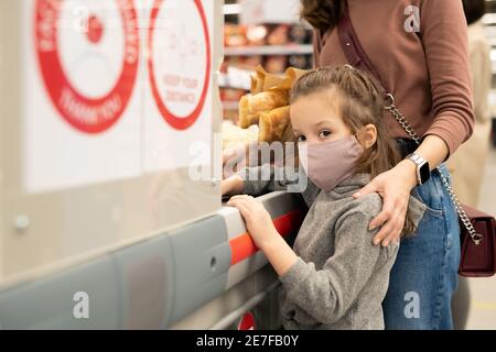 Junge Frau berühren Schulter von ihrem niedlichen kleinen Tochter in casualwear und Schutzmaske beim Stehen am Kassenschalter Supermarkt Stockfoto