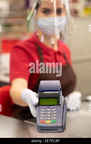 Handschuh der weiblichen Kassiererin in Uniform, Schutzschirm und Maske hält Zahlungsterminal über Kasse im Supermarkt Stockfoto