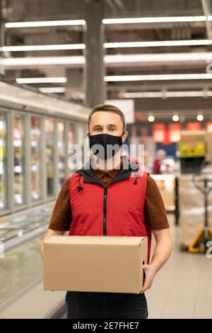 Junger männlicher Kurier in Uniform und Schutzmaske stehen dabei Große Anzeige im Supermarkt und halten verpackte Box mit frischen Produkte Stockfoto