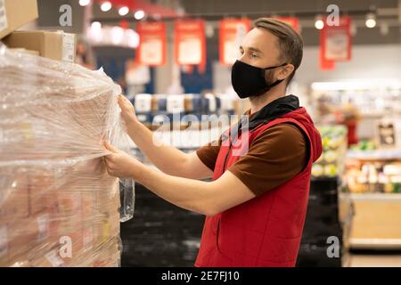 Junger männlicher Arbeiter des modernen Supermarkts in Uniform und schützend Maske Auspacken riesiger Stapel verpackter Kartons mit Waren Stockfoto