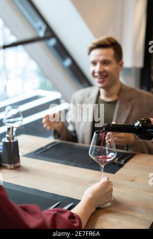 Hand einer jungen Frau, die vor dem Tisch sitzt Von ihrem glücklichen Freund und hält Weinglas während Kellner Gießen Rotwein hinein Stockfoto