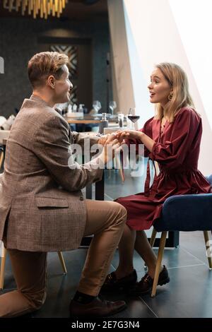 Glücklich jung elegant Mann Blick auf seine überrascht mädchenfieder während Setzen Verlobungsring auf ihren Finger durch serviert Tisch während Abendessen Stockfoto