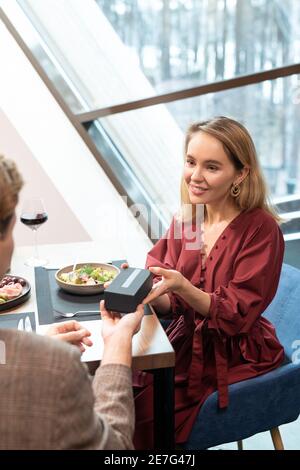 Ziemlich blonde Mädchen vorbei kleine dunkelblaue Box mit Geschenk An ihren Freund, der vor ihr sitzt, serviert Tisch im luxuriösen Restaurant Stockfoto