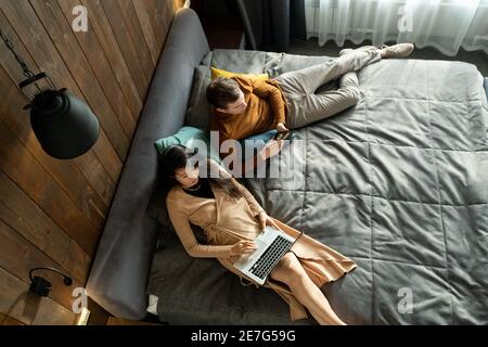 Junge elegante asiatische Frau mit Laptop, während ihr Mann oder Freund Scrollen in Smartphone, beide entspannen auf dem Bett im Hotelzimmer Stockfoto