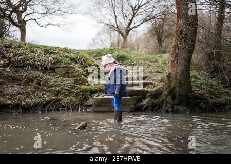 Aufgeregt glücklich junge Mädchen wellinton Stiefel zu Fuß durch die herbstliche Landschaft Flusslauf plantschen im Wasser Erforschen der freien Natur bobble hut Stockfoto