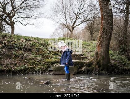 Aufgeregt glücklich junge Mädchen wellinton Stiefel zu Fuß durch die herbstliche Landschaft Flusslauf plantschen im Wasser Erforschen der freien Natur bobble hut Stockfoto