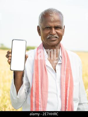 Lächelnder alter indischer Bauer, der vor Ackerland steht Halten Sie das Handy, während Sie in die Kamera schauen Stockfoto