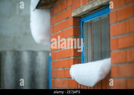 Schneekappen auf der Fensterbank. Eine Wand mit roten Ziegeln und einem blauen Fensterrahmen mit Balken. Stockfoto