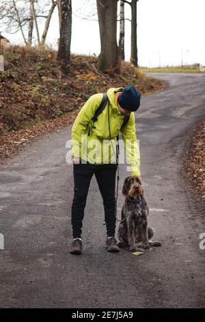 Der junge Mann in einer gelben Jacke kratzt hinter dem Ohr eines Cesky Fousek, eines Jagdhundes, der gehorsam neben seinem Bein sitzt. Binäres Porträt der Aufnahme Stockfoto