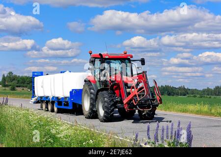 Landwirt fährt Red Case Traktor zieht landwirtschaftlichen Anhänger voller Kunststoff eingewickelten Silage Ballen vom Feld zur Lagerung. Somero, Finnland. Juni 19, 20. Stockfoto
