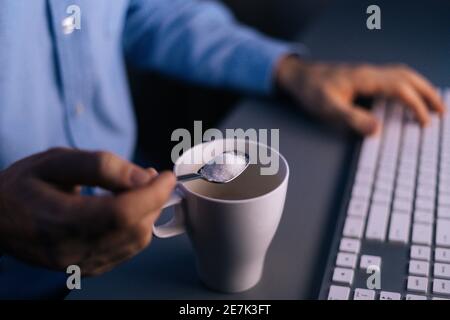 Nahaufnahme der Hände unkenntlich Mann Gießen Zucker auf Kaffee In Tasse mit kleinem Metalllöffel Stockfoto
