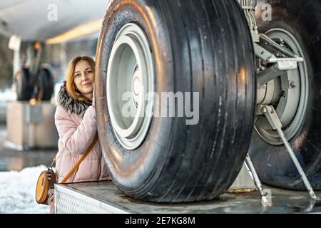 Eine junge Frau schaut hinter großen Rädern heraus. Das vordere Fahrwerk eines Passagierflugzeugs, das am Flughafen repariert wird. Stockfoto