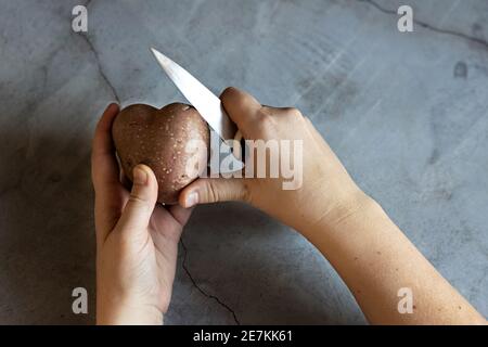 Herzförmige Kartoffeln und ein Küchenmesser in weiblichen Händen auf dem Hintergrund einer grauen Marmorplatte. Essen kochen, Gemüse putzen. Ansicht von einem Stockfoto