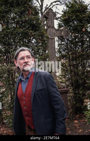 Porträt eines gutaussehenden Mannes in den 50er Jahren, der auf dem Friedhof steht Vor einem Steinkreuz Stockfoto